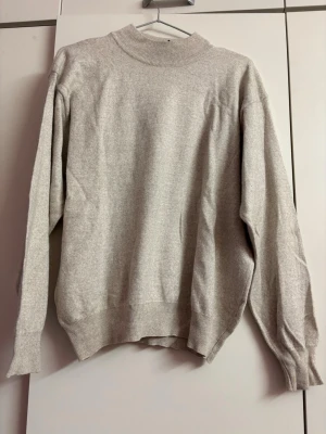 Beige stickad tröja med hög krage - En enkel och stilren beige stickad tröja med hög ribbad krage och långa ärmar. Tröjan har ribbade muddar vid ärmslut och nederkant. Perfekt för lager på lager och passar till det mesta i garderoben.