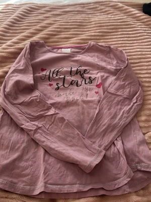 Rosa långärmad topp med text - Söt rosa långärmad topp med trycket 'All the stars make me smile' och små hjärtan på framsidan. Rund halsringning och mjukt material som känns skönt mot huden. Perfekt för dig som gillar gulliga detaljer och bekväma plagg.