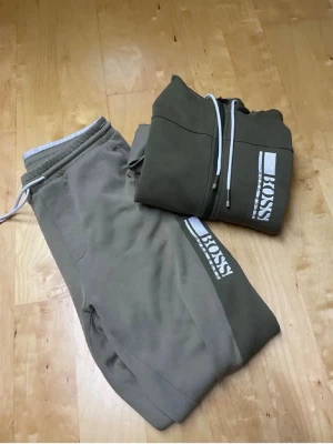 Två BOSS hoodies och byxor set - Säljer två snygga set från BOSS med hoodie och matchande byxor. Ena setet är olivgrönt med vit logga och dragkedja, det andra är svart med gulddetaljer och dragkedja. Perfekt för en chill och stilren look. Mjukt material och bekväm passform.