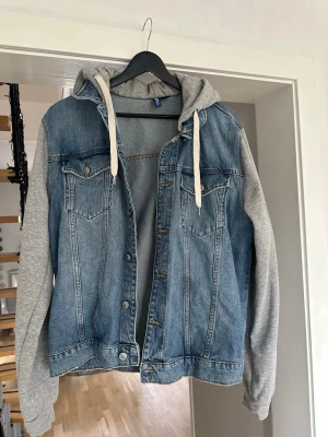 Blå jeansjacka med grå luvärm - Snygg blå jeansjacka från H&M med grå sweatshirt-ärmar och huva. Jackan har två bröstfickor med lock och knappar samt dragsko i luvan. Perfekt för en avslappnad streetstil.
