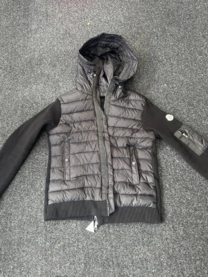 Svart cardigan med huva från Moncler - Snygg svart cardigan från Moncler med stickade ärmar och ribbade muddar. Jackan har två fickor framtill med tryckknappar, dragkedja och en huva. Moncler-logga på ärmen och coolt serietidningsfoder på insidan. Storleken är egentligen M men men passar som S