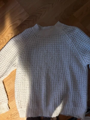 Vit grovstickad tröja - Säljer en vit grovstickad tröja med hög ribbad krage och långa ärmar. Tröjan har ett luftigt, strukturerat mönster och ribbade muddar vid ärmslut och nederkant. Perfekt för lager på lager och en avslappnad stil.