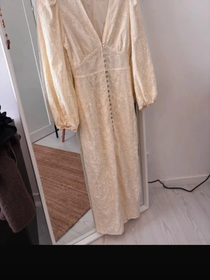 Långklänning i spets från Adoore - Elegant långklänning från Adoore i ljus champagnefärg med v-ringning, långa puffärmar och fina spetsdetaljer. OBS, klänningen har justerats hos skräddare. Kortat ner 10cm på längden samt kortat ner armarna både på bredd/längd (se sista bilder för mått). Endast använd 1 găng! I en ljusgul/krämvit färg.