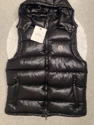 Moncler väst - Snygg svart väst med en luva från moncler. Passar dig som gillar en skön stil!