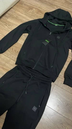 Svart loungewear-set från BOSS - Snyggt svart loungewear-set från BOSS med dragkedja på hoodien och matchande byxor med dragsko i midjan. Setet har diskreta BOSS-loggor på både tröja och byxa samt praktiska fickor med dragkedja. Perfekt för chill dagar hemma.