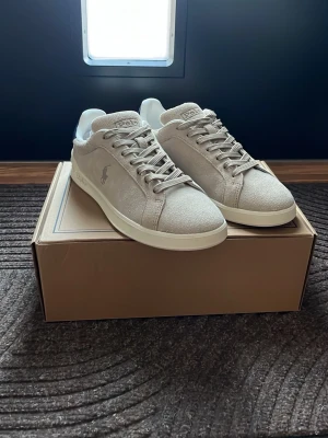 Beige sneakers från Polo Ralph Lauren - Stilrena beige sneakers från Polo Ralph Lauren i mocka med vita sulor och mörkblå detalj på hälen. Klassisk låg modell med snörning och diskret logga på sidan. Perfekta för en clean och avslappnad look. Skorna är sprillans nya och nypris ligger på 1695kr. Mitt pris 1000kr