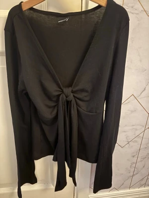Svart långärmad topp med knyt - Trendig svart långärmad topp med djup v-ringning och knyt framtill. Tunn och mjuk i materialet, perfekt att styla lager på lager eller som den är. Enkel men snygg design som passar till många olika looks.