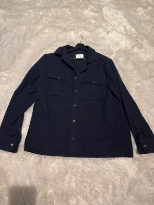 Mörkblå overshirt från BROTHERS - Stilren mörkblå overshirt från Brothers med klassisk krage och två stora bröstfickor med lock. Skjortan har raka ärmar med manschett och knappar framtill. Perfekt att slänga över en t-shirt för en avslappnad look.