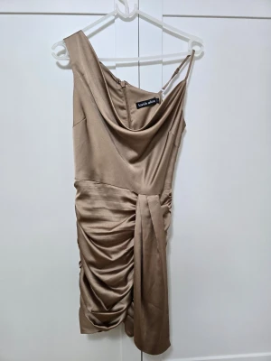 Champagnefärgad asymmetrisk klänning  - Snygg kort klänning i champagnefärgad satin från Lavish Alice. Klänningen har asymmetrisk axel, draperad framsida och en slits. Materialet är glansigt och känns lyxigt. 