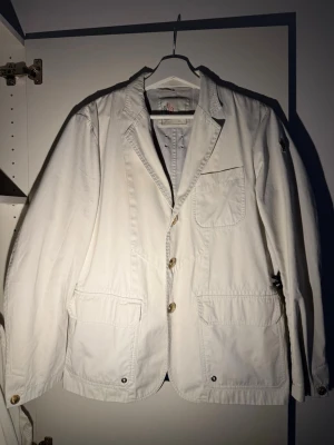 Vit kavaj från Moncler - Stilren vit kavaj från Moncler med tre knappar framtill, tre utanpåliggande fickor och klassisk krage. Tillverkad i bomullstyg med avslappnad passform och diskret Moncler-logga på ärmen. Perfekt för en trendig och avslappnad look. Se liten reva på sista bilden, materialet är såpass tätt så den inte syns vid användning 