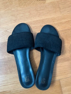 Svarta sandaler från XIT - Stilrena svarta sandaler från XIT med bred ribbad rem över foten och platt sula. Ovandelen är i textil och sulan i syntetmaterial. Perfekta för en avslappnad och trendig look.