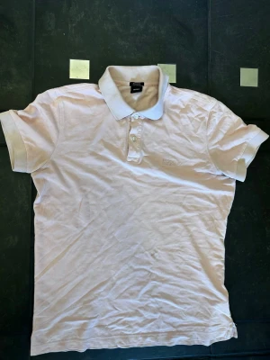 Rosa pikétröja från Hugo Boss - Klassisk rosa pikétröja från Hugo Boss i mjuk pima bomull. Tröjan har korta ärmar, krage med knappar och diskret Boss-logga på bröstet. Regular fit för en avslappnad men snygg look.