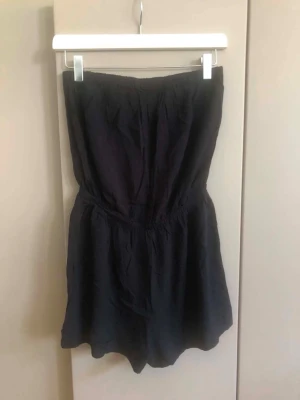 Svart tubjumpsuit med shorts - Säljer en svart, axelbandslös jumpsuit med shorts och resår i midjan. Perfekt för sommaren och enkel att styla med sneakers eller sandaler. Materialet känns lätt och luftigt, vilket gör den skön att bära under varma dagar.