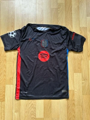 Svart FC Barcelona matchtröja Nike - Snygg svart FC Barcelona matchtröja från Nike med röda och blå detaljer på sidorna. Tryck med Spotify-logga, Champions League-märke på ärmen och Lamine Yamal 19 på ryggen. Tillverkad i lätt och ventilerande material, perfekt för fotboll eller träning.