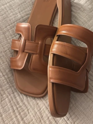 Bruna slip-in sandaler med bred rem - Snygga bruna slip-in sandaler med breda remmar i konstläder och fyrkantig tå. Stilsäker design med utstansade detaljer på ovandelen. Perfekta för en trendig och avslappnad look till sommaren.