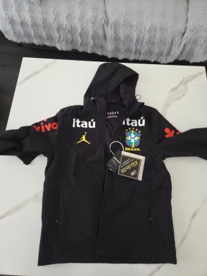 Svart Brasilien Vindjacka Jordan  - Snygg svart vindjacka från Jordan med Brasilien CBF-logga, gula och vita tryck samt röda detaljer på ärmarna. Jackan har huva, dragkedja och är tillverkad i GORE-TEX-material som står emot regn och vind. Perfekt för dig som gillar sportig stil och vill sticka ut.