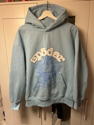 Ljusblå SP5DER hoodie - Ljusblå hoodie från Sp5der med stor huva och magficka. På bröstet finns ett stort tryck med ett blått spindelnät och texten 'sp5der' i vit, fluffig stil. Hoodien har ribbade muddar vid ärmar och nederkant. Perfekt för dig som gillar streetwear och unika prints. Storlek M.