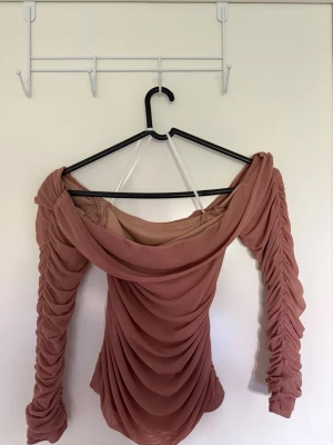 Rosa offshoulder topp med drapering - Trendig offshoulder topp i ljusrosa med snygg drapering framtill och längs ärmarna. Toppens material är stretchigt och tunt, vilket ger en smickrande passform. Perfekt för dig som gillar en feminin och modern stil.