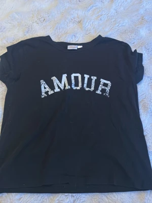 Svart t-shirt med AMOUR från Gina Tricot - Cool svart t-shirt från Gina Tricot med texten 'AMOUR' i vit, lite sliten print över bröstet. Klassisk rund halsringning och korta ärmar. Perfekt till jeans eller kjol för en avslappnad look.