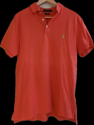 Korallröd pikétröja från Ralph Lauren - Snygg korallröd pikétröja från Ralph Lauren med klassisk krage, korta ärmar och två knappar framtill. Ikoniska gröna loggan broderad på bröstet. Perfekt till sommaren och enkel att matcha med jeans eller shorts.