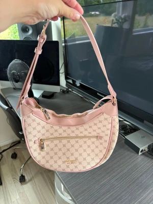 Ljusrosa axelväska från Guess 💗 - Snygg ljusrosa axelväska från Guess med justerbart axelband och guldfärgade detaljer. Väskan har ett mönstrat tyg med Guess-loggor, dragkedja upptill och en praktisk ytterficka med dragkedja framtill. Perfekt för dig som gillar trendiga accessoarer.