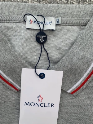 Grå Moncler pikétröja med röd detalj - Snygg grå pikétröja från Moncler med klassisk krage och knappar framtill. Kragen har vita och röda ränder för en sportig touch och bröstet pryds av Monclers ikoniska logotyp. Perfekt för en clean och stilren look.