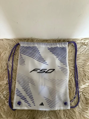 Vit Adidas F50 gympåse - Säljer en vit gympåse från Adidas med F50-tryck och lila detaljer. Påsen har dragsko i lila snöre och grafiskt mönster i lila och svart. Perfekt för träning eller att bära med sig det viktigaste. Lätt och smidig design i syntetmaterial.