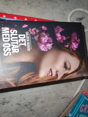 Det slutar med oss av Colleen Hoover - En gripande roman med svart omslag och rosa blommor, skriven av Colleen Hoover. Boken handlar om kärlek, svåra val och att hitta sig själv. Perfekt för dig som gillar starka berättelser och känslor. Omslaget har ett porträtt av en kvinna med blommor.