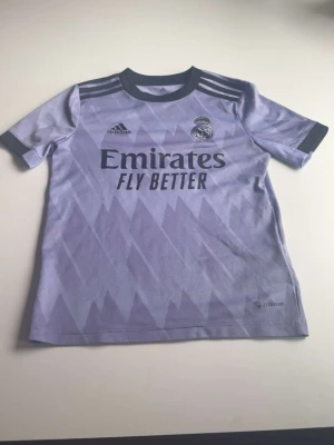 Lila Real Madrid matchtröja Adidas - Säljer en lila Real Madrid matchtröja från Adidas med mörklila detaljer och grafiskt mönster. Tröjan har klubbmärke, Adidas-logga och sponsortryck på bröstet. Korta ärmar och rund hals. Perfekt för fotbollsträning eller att bära som supporter.