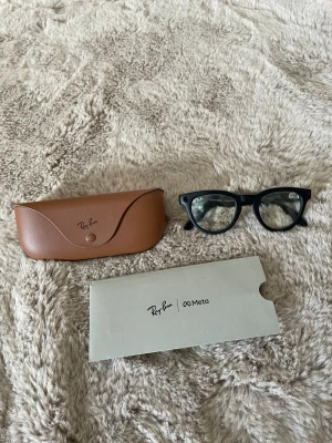  Ray-Ban Meta  - Säljer ett par svarta Ray-Ban Meta Wayfarer-solglasögon med klassisk form. Bågarna är i svart syntetmaterial och kommer med ett brunt fodral i skinn. Perfekta för dig som vill ha en tidlös och ikonisk look.