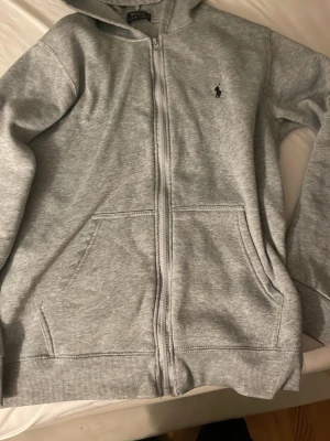 Grå zip hoodie från Polo Ralph Lauren - Snygg grå hoodie med dragkedja från Polo Ralph Lauren. Klassisk design med huva, två fickor framtill och den ikoniska lilla loggan broderad på bröstet. Tillverkad i mjukt sweatshirtmaterial som är perfekt för chill dagar.