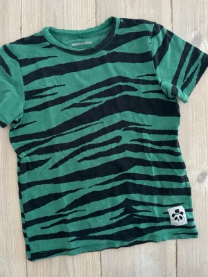 Grön zebramönstrad t-shirt Mini Rodini, stl 104/110 (4-5 år) - Cool grön t-shirt från Mini Rodini med svart zebramönster och en liten panda-patch nertill. T-shirten har rund hals och korta ärmar, perfekt för en avslappnad och trendig stil.