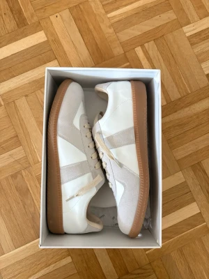 Vita maison margiela gats storlek 44 - Snygga sneakers i vitt och beige med klassisk retrostil. Ovandelen är i en mix av slätt läder och mocka, och sulan är i brunt gummi för en clean look. Skorna har vita snören och diskreta detaljer som gör dem lätta att matcha till olika outfits.