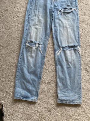 Ljusblå ripped jeans från Gina tricot  - Säljer ett par ljusblå jeans från Perfect Jeans med raka ben och flera slitna detaljer och hål på framsidan. Klassisk femficksmodell med dragkedja och knapp. Perfekta för en avslappnad och trendig look.