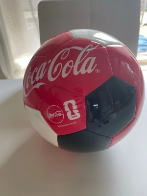 Coca-Cola x FIFA fotboll - Snygg fotboll i samarbete mellan Coca-Cola och FIFA. Bollen har klassiskt mönster i rött, svart och vitt med Coca-Cola-logga tryckt på flera paneler. Tillverkad i slitstarkt syntetmaterial, perfekt för spel på gräs eller konstgräs.