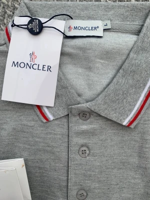 Grå Moncler pikétröja med röd/vit kant - Snygg grå pikétröja från Moncler med klassisk krage och knäppning framtill. Kragen har stilrena röda och vita ränder och bröstet pryds av Monclers ikoniska logotyp. Tillverkad i mjukt bomullsmaterial som känns skönt mot huden.