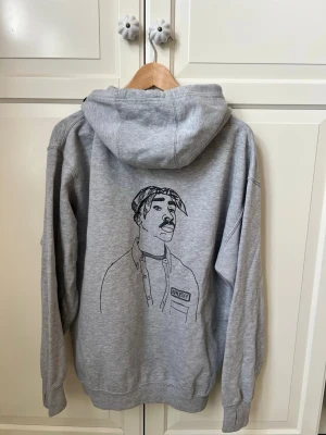 Grå hoodie med tryck från Appetiff - Säljer en grå hoodie från Appetiff med en stor svart linjeteckning av en person på ryggen. Hoodien har huva och ribbade muddar vid ärmslut och nederkant. Tillverkad i mjukt sweatshirtmaterial, perfekt för en avslappnad streetwear-look.