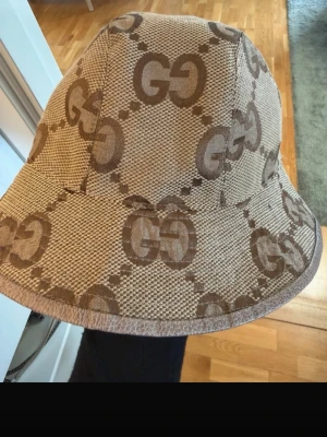 Beige bucket hat från Gucci - Säljer en beige bucket hat från Gucci med det klassiska GG-mönstret i brunt. Hatten är tillverkad i ett slitstarkt tyg och har en bred, mjuk brätte. Perfekt för dig som vill ha en trendig accessoar med lyxig känsla och tydlig logga.