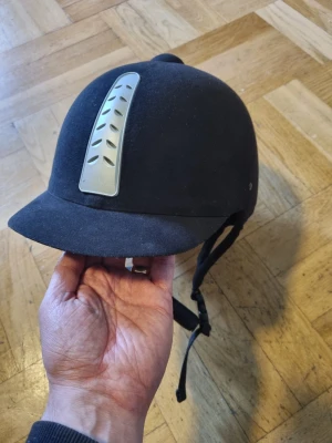 Svart ridhjälm med silverventil - Stilren svart ridhjälm med matt finish och en snygg silverfärgad ventilationsdetalj framtill. Sparsamt använd i bra skick. 