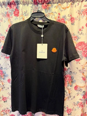 Svart Moncler t-shirt med patch - Snygg svart t-shirt från Moncler med klassisk rund hals och korta ärmar. På bröstet sitter en orange Moncler-patch som ger en clean men exklusiv vibe. Tillverkad i mjuk bomull för skön känsla hela dagen.