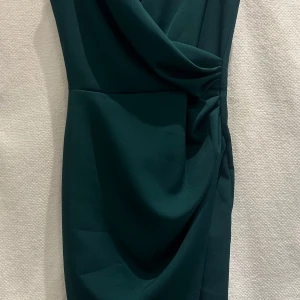 Grön festklänning  - Vacker mörkgrön klänning av märket Katniss.  Figursydd med slits. Dragkedja i ryggen. Bekvämt material.   Strl: 36 men lite åt det mindre hållet. 