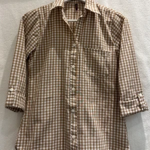 Brunrutig skjorta - Rutig skjorta 3/4 ärm som sätts upp med knapp.   Strl: S men lite åt det mindre hållet, se mått på bild. 