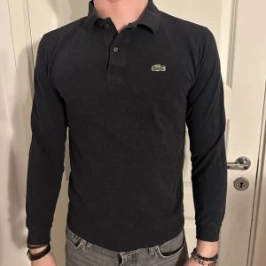 Grå LACOSTE Polotröja/Pikétröja - Stilren Långärmad Grå LACOSTE Polotröja/Pikétröja i extremt bra skick! Storlek M men passar även som S. Modellen är 182 cm och väger 75 kg!