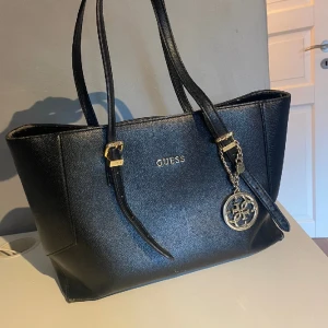 Guess väska  - Säljer denna Guess väska då den inte kommer till användning längre🖤🖤 Har små tecken på användning men fortfarande i fint skick. Är justerbar i banden, hängsmycket utanpå är avtagningsbart, den innehåller flera fack inuti. Skriv privat för fler bilder