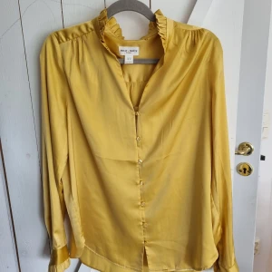 Gul långärmad blus - Senapsgul blus med volangdetaljer vid krage och ärmarna. Varsamt använd, i bra skick. Finns en liten liten fläck som går bort i tvätten. 
