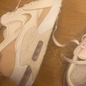 Ett par Nike Air Max sneakers i beige,vit och rosa färg. Skorna har en synlig luftdämpning i sulan och är dekorerade med Nike-logotypen på sidan och hälen. De har snörning framtill och är tillverkade i syntetmaterial.