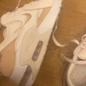 Nike Air Max sneakers i beige och vit - Ett par Nike Air Max sneakers i beige,vit och rosa färg. Skorna har en synlig luftdämpning i sulan och är dekorerade med Nike-logotypen på sidan och hälen. De har snörning framtill och är tillverkade i syntetmaterial.
