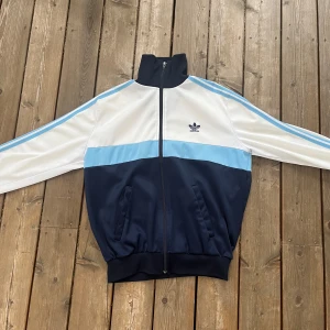 Vintage adidasjacka - En blå och vit vintage adidasjacka från 70-talet i storlek s/m och skulle säga att den sitter lagom oversized på en person som brukar ha på sig kläder i storlek s. Det enda tydliga tecken på användning är ett litet hål på ryggen (se sista bilden)