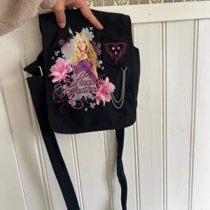 En svart axelväska med ett färgglatt motiv av Hannah Montana. Väskan har rosa och vita blommor samt en kedjedetalj på framsidan. Den har en justerbar axelrem och en klaff med tryckknapp för stängning.