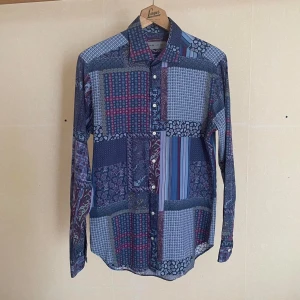 Mönstrad skjorta från Etro - Etro- En långärmad skjorta  med en klassisk  stilig krage och resten består av ett unikt patchwork-mönster där paisley mönstret, som är kännemärket för det italienska exklusiva designerhuset, syns ”här och där”. Skjortan innehåller även en häftig sammansättning av färg i olika nyanser av blått, rött och lila.  -Storlek: 40eu= Medium  -Material: 100% bomull, en mycket mjuk och len typ av bomull -Mått: Längd 74,5 Axlar 44 cm Byst 52 cm Midja 48 cm Ärm 51 cm -Skick: Mycket bra, ”begagnad” är ett val som stavas brist på bevis, då skjortan saknar ”yttre tecken” på användning.